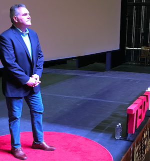 Jody Wood on stage giving a TedX talk.  He is wearing a blue suit jacket, a light blue dress shirt, jeans and brown dress shoes, and looks out at his audience with his hands clasped in front of his waist.