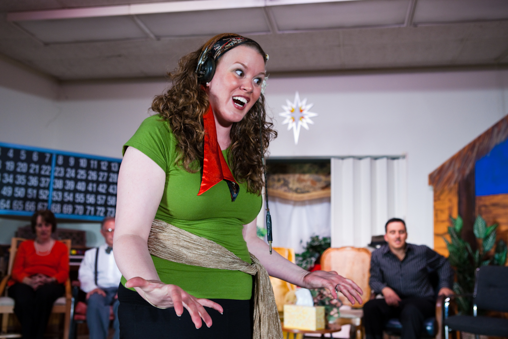 Stephanie Overton-Hall wears a green shirt with black pants, and a gold scarf tied around her waist as a makeshift belt.  She wears a colorful paisley scarf tied around her head as a headband, and also has large black headphones on her ears.  She stands in the front of a room with several other people in chairs in the background, out of focus.  She has her hands outstretched, palms up, with a wild and exasperated look on her face.