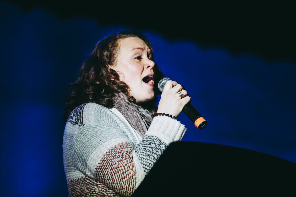 Stephanie Overton-Hall is wearing a woven striped cardigan and her hair is brown and curly. She holds a microphone and sings passionately against a dark black background to her invisible audience.