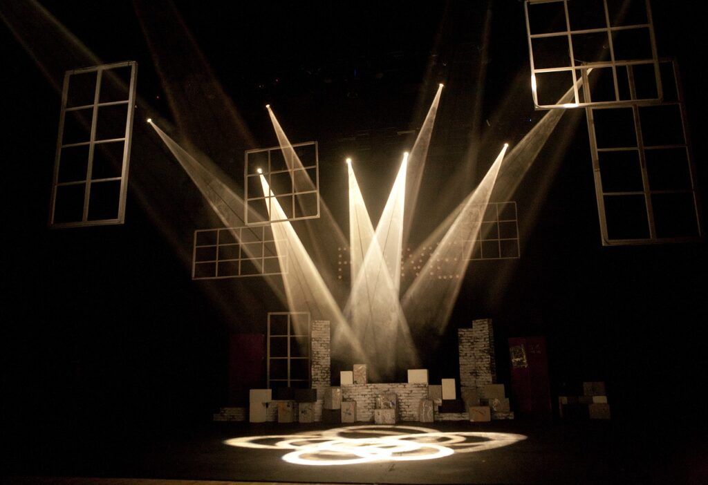 A black and white image of a theatre with lights streaming from all directions to a single point on the stage.